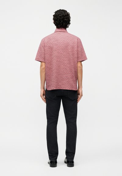 Άνδρας με σγουρά μαλλιά, ντυμένος με κοντομάνικο κόκκινο πουκάμισο με σχέδια, μαύρο τζιν και μαύρα παπούτσια, στέκεται με την πλάτη γυρισμένη σε λευκό φόντο.