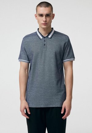 Młody mężczyzna w szarej koszulce polo z krótkim rękawem, z białym i granatowym paskowanym kołnierzykiem oraz mankietami, stoi na jednolitym tle.