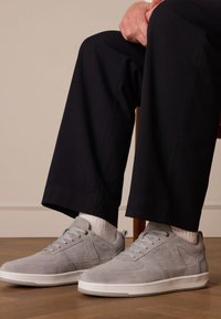 Grijze suède sneakers met witte rubberen zolen, voorzien van een vetersluiting en textuuraccenten, gedragen met donkere broeken en witte sokken.