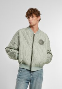 Giacca bomber trapuntata verde chiaro con zip frontale, polsini e bordo a coste, e una toppa ricamata rotonda sul lato sinistro del petto.
