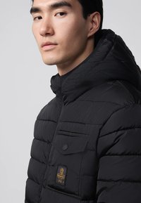 Giacca imbottita nera con cappuccio, caratterizzata da cuciture orizzontali, una tasca frontale con chiusura a pressione e una patch con logo sul petto.