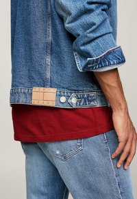 Chaqueta de mezclilla con un lavado azul claro, que presenta una etiqueta de cuero con "TOMMY JEANS", botones de metal y costuras rojas en los puños. Camisa roja debajo.