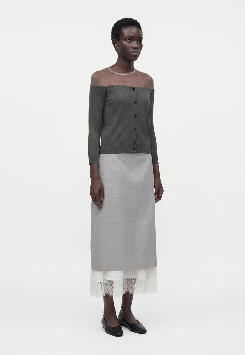 Woman wearing an off-shoulder buttoned dark gray top, long gray skirt with white lace hem, and black pointed flats, standing against white background.