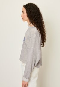 Sivi dolg rokav srajc z ohlapnim krojem, rebrastimi manžetami in majhnim modrim logotipom na prsih, v kombinaciji z belimi kavbojkami.