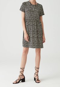 Schwarzes, kurzärmliges Kleid mit floralem Muster und gerafftem Bund, verziert mit kleinen weißen und gelben Blumen. Kombiniert mit schwarzen, gebrochenen Sandalen mit Absatz.