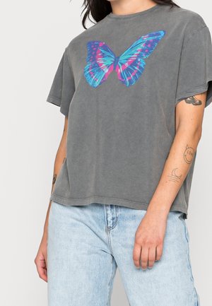 Osoba nosząca wyblakły szary t-shirt z dużą niebiesko-różową grafiką motyla, jasnoniebieskie dżinsy i widoczne tatuaże na rękach.