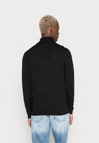Maglione nero a maglia con colletto alto, maniche lunghe e texture a coste. Indossato con jeans chiari e sbiaditi, che mostrano un outfit casual.