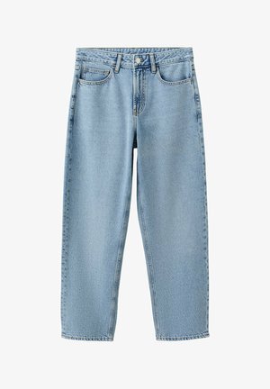 Jeune homme portant un t-shirt bleu marine, un jean bleu clair et des chaussures marron, debout et appuyé contre un mur blanc.
