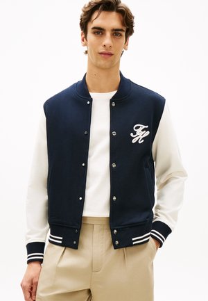 Young man wearing a navy varsity jacket with white sleeves, a white shirt, and beige pants, standing with one hand in pocket.