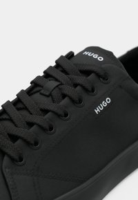 Μαύρα καμβά παπούτσια που διαθέτουν επίπεδες μαύρες κορδόνες, λείο υφαντό φινίρισμα και λευκή επωνυμία "HUGO" κοντά στα τρυπήματα.