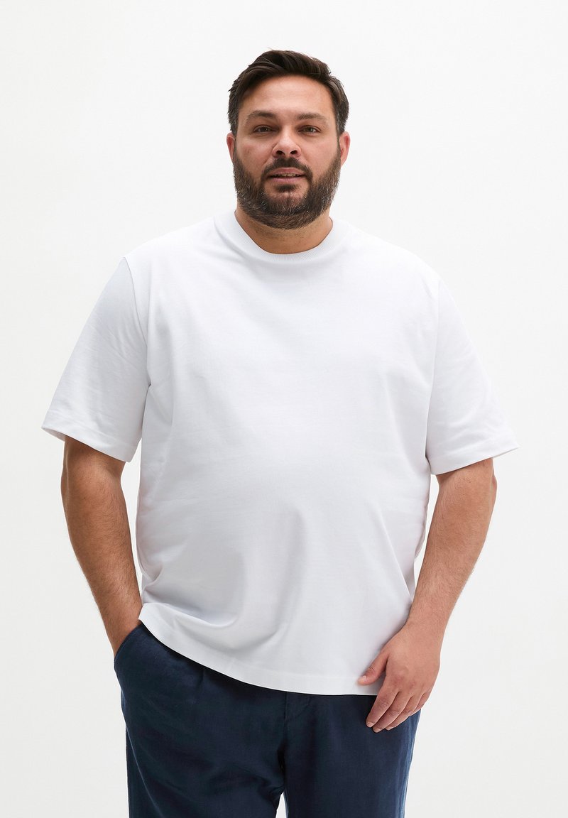 Homme barbe portant un t-shirt blanc uni à manches courtes et un pantalon bleu marine, debout devant un fond blanc avec une main dans la poche.