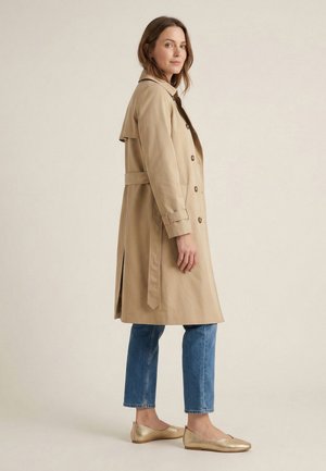 Kobieta stojąca, ubrana w beżowy trencz na niebieskich dżinsach i złote baleriny, zwrócona bokiem na jednolitym tle.