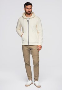 Felpa con cappuccio crema con zip frontale, abbinata a pantaloni cargo beige. Sneakers bianche completano l'outfit. Design semplice e casual.
