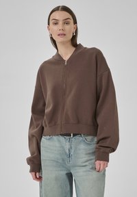 Brown cropped sweatshirt with a zip closure, ribbed cuffs, and a relaxed fit. Paired with light blue loose-fitting jeans.
