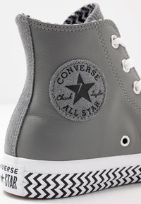 Converse Chuck Taylor montantes grises avec un patch logo en daim noir, des œillets blancs et une semelle en caoutchouc à motif zigzag.