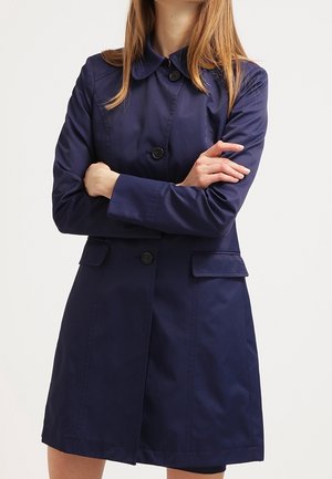 Femme portant un trench-coat bleu marine avec des boutons et des poches à rabat, debout, les bras croisés, devant un fond uni.