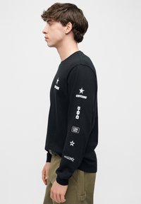 Νεαρός άνδρας με μαύρο πουκάμισο μακρυμάνικο με πολλαπλά λευκά λογότυπα Converse στο στήθος και το μανίκι, φορώντας χακί παντελόνια, κοιτάζει προς τα αριστερά.