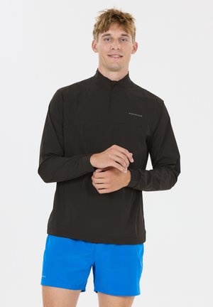 Ung mand tilpasser håndled, iført sort langærmet trøje med lynlås og lyseblå atletiske shorts, stående mod en ensartet hvid baggrund.