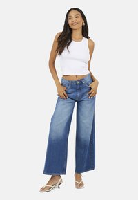 Top corto bianco a coste leggero abbinato a jeans blu in denim a gamba larga, con vita alta e finitura scolorita. Indossato con sandali bianchi.
