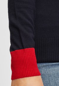 Zbliżenie na rękaw granatowego swetra z jasnoczerwonym ściągaczem ponad jasnoniebieskimi dżinsami.