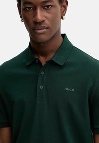 Polo de color verde oscuro con cuello, dos botones y un sutil logo "HUGO". La tela parece texturizada y duradera.