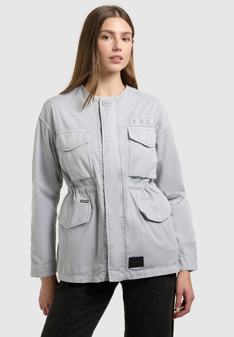 Femme aux cheveux raides portant une veste boutonnée gris clair avec quatre poches à rabat et un pantalon noir, sur un fond uni.
