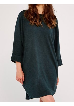 Kobieta ubrana w ciemnozieloną, oversize'ową dzianinową sukienkę z rękawami podwiniętymi do ćwiartek, stojąca na jednolitym jasnym tle.