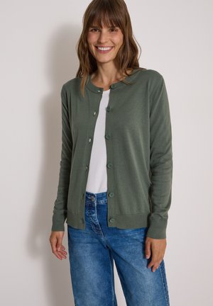 Femme souriante aux cheveux bruns portant un cardigan vert boutonné sur un haut blanc et un jean bleu sur fond clair uni.