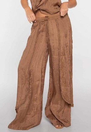 Pantalones marrones arrugados de pierna ancha con cintura de cordón y paneles de tela superpuestos estilo envolvente, usados por una persona de pie.