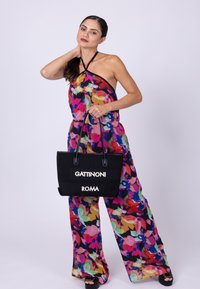 Svart tote-väska med "GATTINONI" och "ROMA" i vit text. Modellen bär en livfull, blommönstrad jumpsuit i rosa, gröna och gula nyanser.