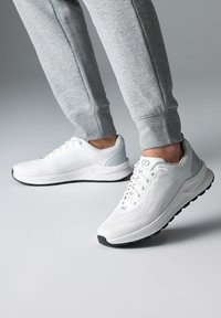 Weiße Sportschuhe mit strukturiertem Mesh-Obermaterial, hellgrauen Akzenten und schwarzer Gummisohle. Tragen mit grauen Jogginghosen.