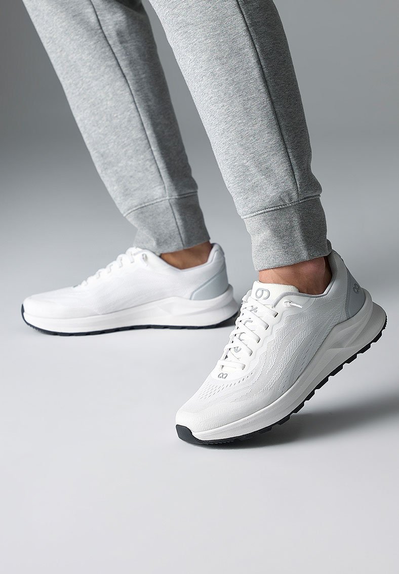Weiße Sportschuhe mit strukturiertem Mesh-Obermaterial, hellgrauen Akzenten und schwarzer Gummisohle. Tragen mit grauen Jogginghosen.
