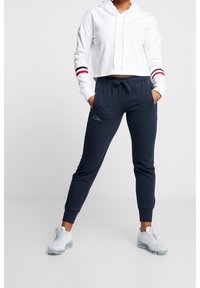 Vit kortärmad hoodie med röda och marinblå randiga detaljer, kombinerad med marinblå joggers med en broderad logotyp, buren med ljusa sneakers.