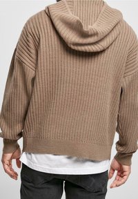 Stickad, brun hoodie med ribbad textur och en avslappnad passform. Har en huva och sänkta axelsömmar, lagrad över en vit skjorta.