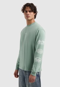 Mintgroene T-shirt met lange mouwen van zacht katoen. Heeft witte grafische prints op beide mouwen met logo's en tekst. Comfortabele pasvorm, ronde hals.