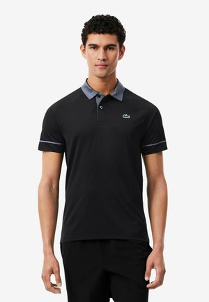 Giovane uomo che indossa una polo nera con colletto grigio e piccolo logo coccodrillo sul petto sinistro, in piedi davanti a uno sfondo bianco.