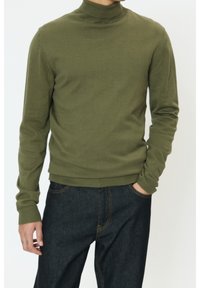 Pull col roulé vert olive en tissu doux et texturé, manches longues avec poignets côtelés, associé à un bas en denim foncé.