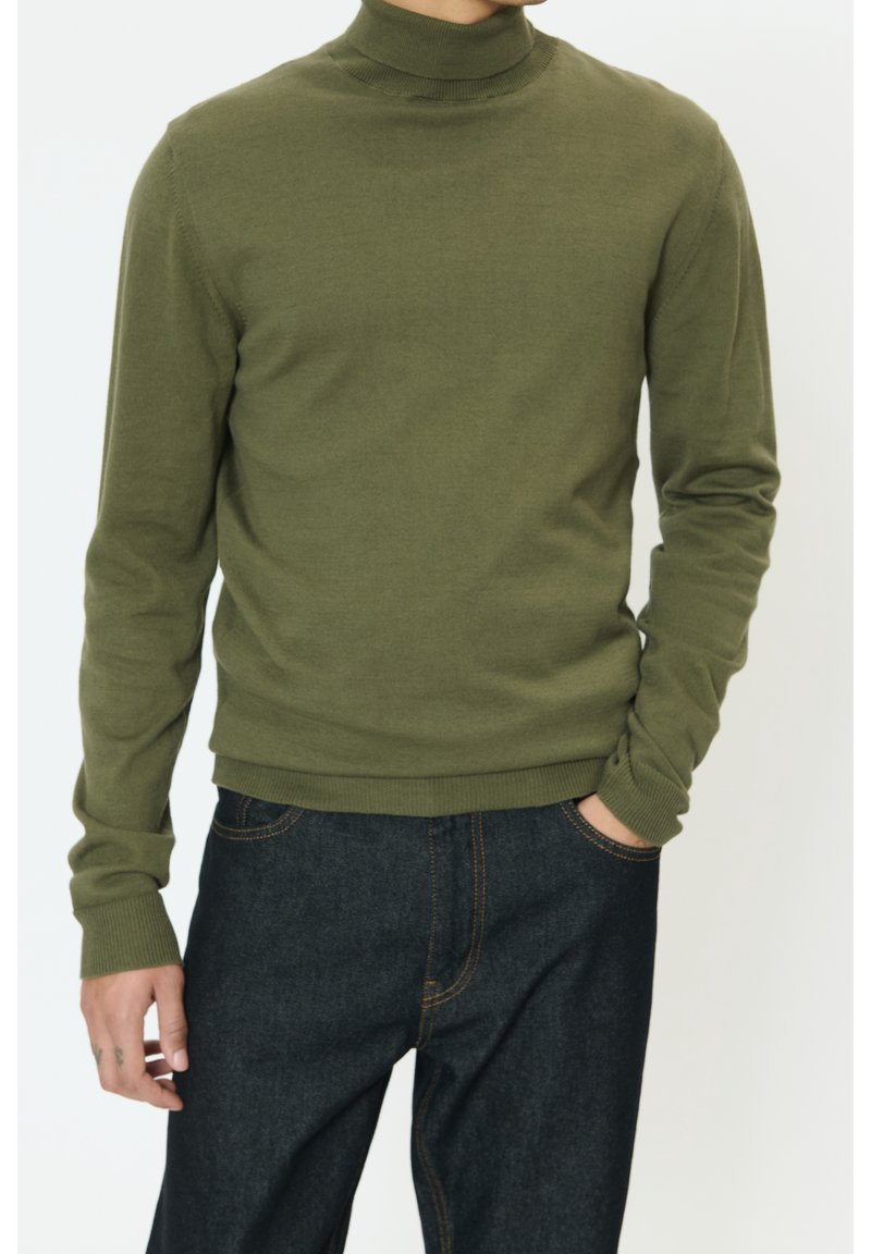 Pull col roulé vert olive en tissu doux et texturé, manches longues avec poignets côtelés, associé à un bas en denim foncé.