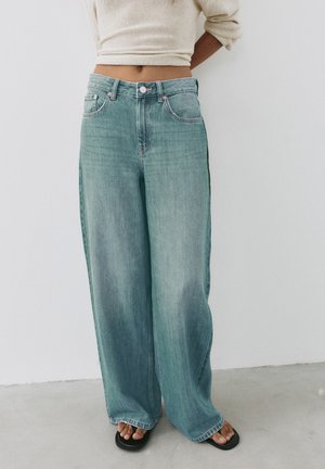 Person iført lyseblå vidde bukser, beige cropped sweater og sorte sandaler med åben tå, stående mod en hvid væg.