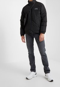 Doudoune noire avec col montant, poignets élastiques et deux poches avant. Portée avec un jean foncé et des baskets blanches.