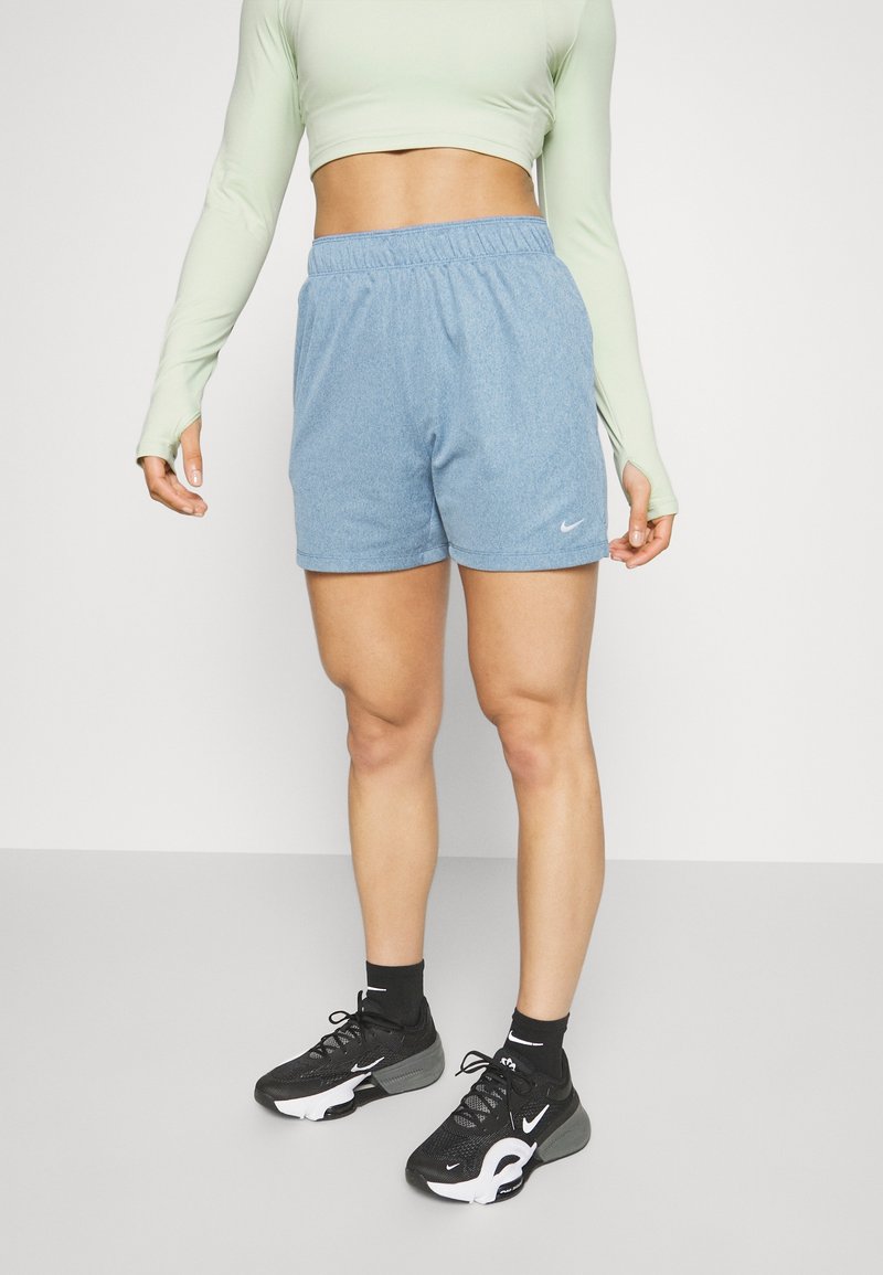 Femme portant un crop top manches longues vert clair, un short bleu clair à taille élastique, des chaussettes noires et des chaussures de sport noires, debout sur un sol gris.