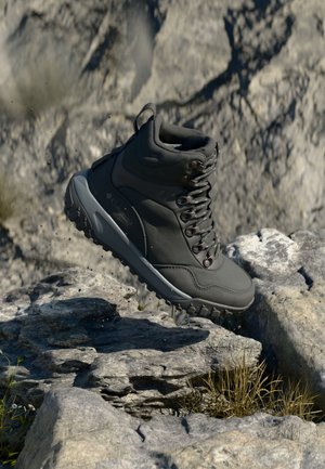 Botte de randonnée grise foncée en l'air au-dessus d'un terrain rocheux avec des particules de terre visibles, mettant en valeur la semelle robuste et le design à lacets.