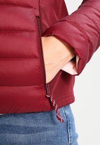 Burgundin värinen puhallettava takki, jossa on tikattu pinta, vetoketjullinen tasku ja säädettävä nauhakuomu, yhdistettynä farkkuihin.