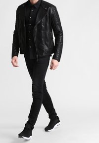 Zwarte leren jas met doorgestikte schouders, gecombineerd met een donker shirt, slim-fit zwarte jeans en zwarte sneakers met witte accenten.