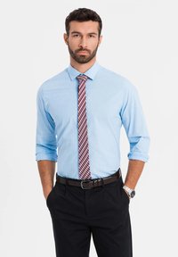 Camicia button-up azzurro chiaro con maniche arrotolate, abbinata a una cravatta a quadri rossa e blu. Pantaloni neri e una cintura marrone con un orologio argentato.