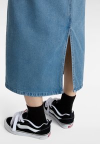 Jambes portant des chaussettes noires et des baskets noir et blanc, partiellement couvertes par une jupe en jean bleue avec une fente au dos.