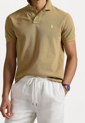 Polo à manches courtes kaki avec col et deux boutons, présentant un petit logo brodé sur la poitrine gauche, porté avec un short en lin blanc.