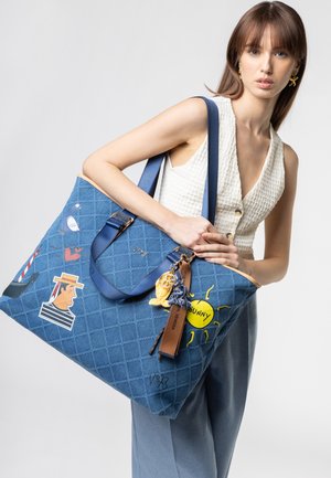Mujer con top blanco sin mangas y pantalones azules que sostiene un gran bolso tote azul acolchado con parches de colores y accesorios de llavero.