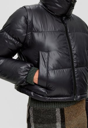 Persoon die een zwarte glanzende donzen jas draagt met rits en handen in de zak, gecombineerd met een groene trui en een bruine geruite rok.