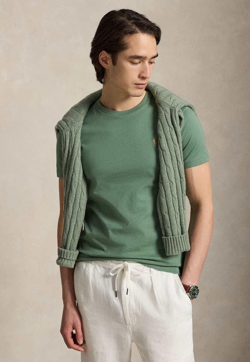 Mlajši moški v zeleni majici s puloverjem s kitami, oblečen v bele hlače z vrvico in z ročno uro.